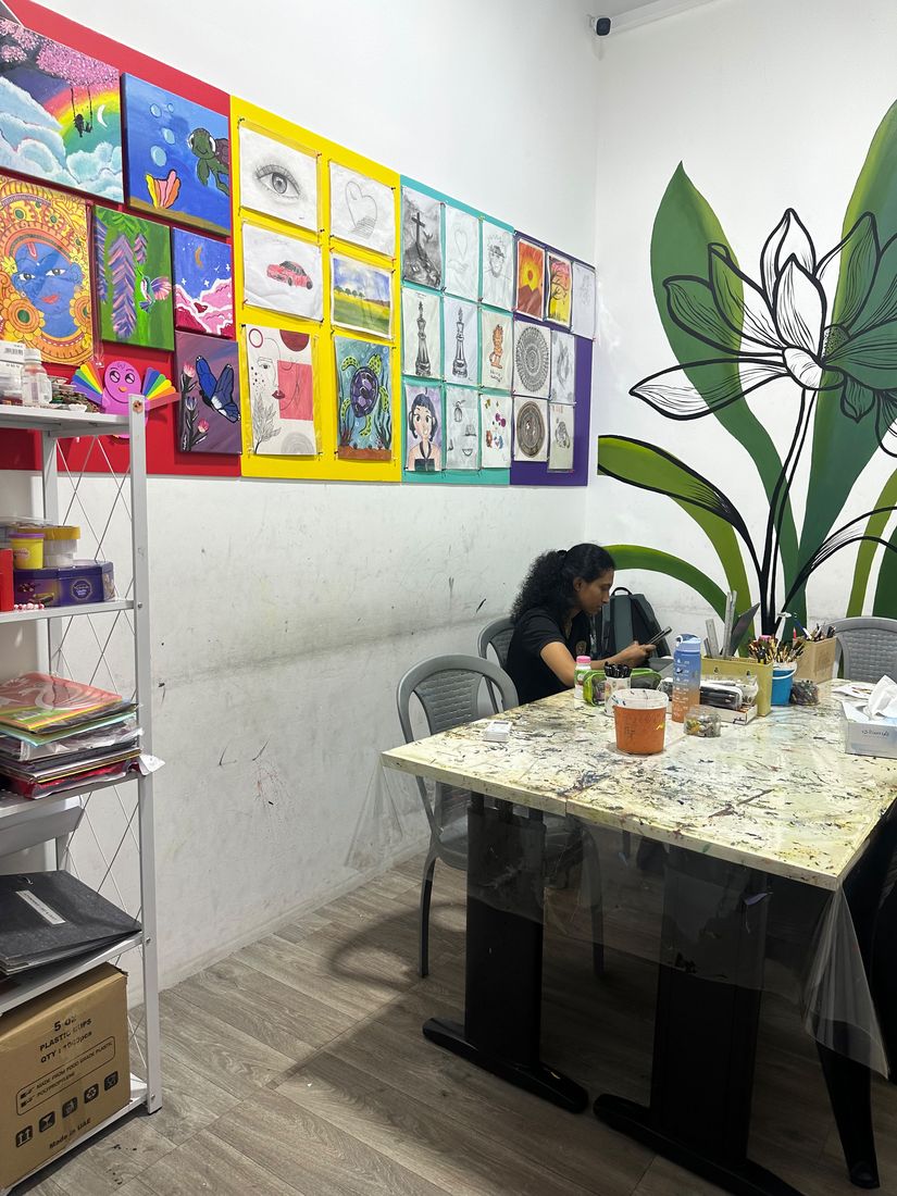 Art room wall covered in student paintings and drawings, with a student at the worktable